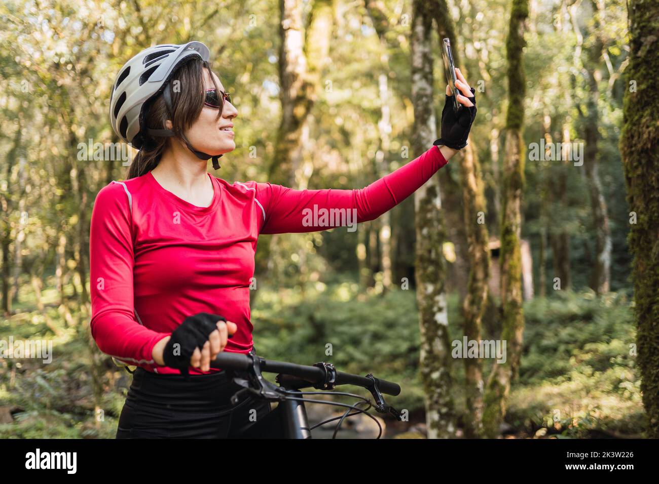Lächelnde Biker-Frau im Helm sitzt auf dem Fahrrad, während sie auf dem Handy im Wald Selbstporträt aufnahm Stockfoto