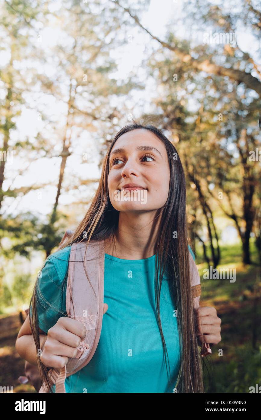 Selbstvertrauen. Junge Frau mit türkisfarbenem T-Shirt, Rucksack mit gelassenem Lächeln im Wald. Vertikal Stockfoto