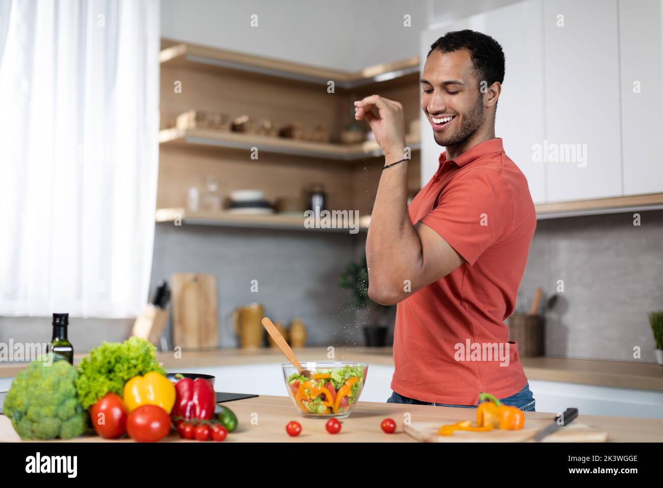 Happy lustige Millennial african american Kerl Salze Salat, bereiten Sie Essen am Tisch mit Bio-Gemüse Stockfoto