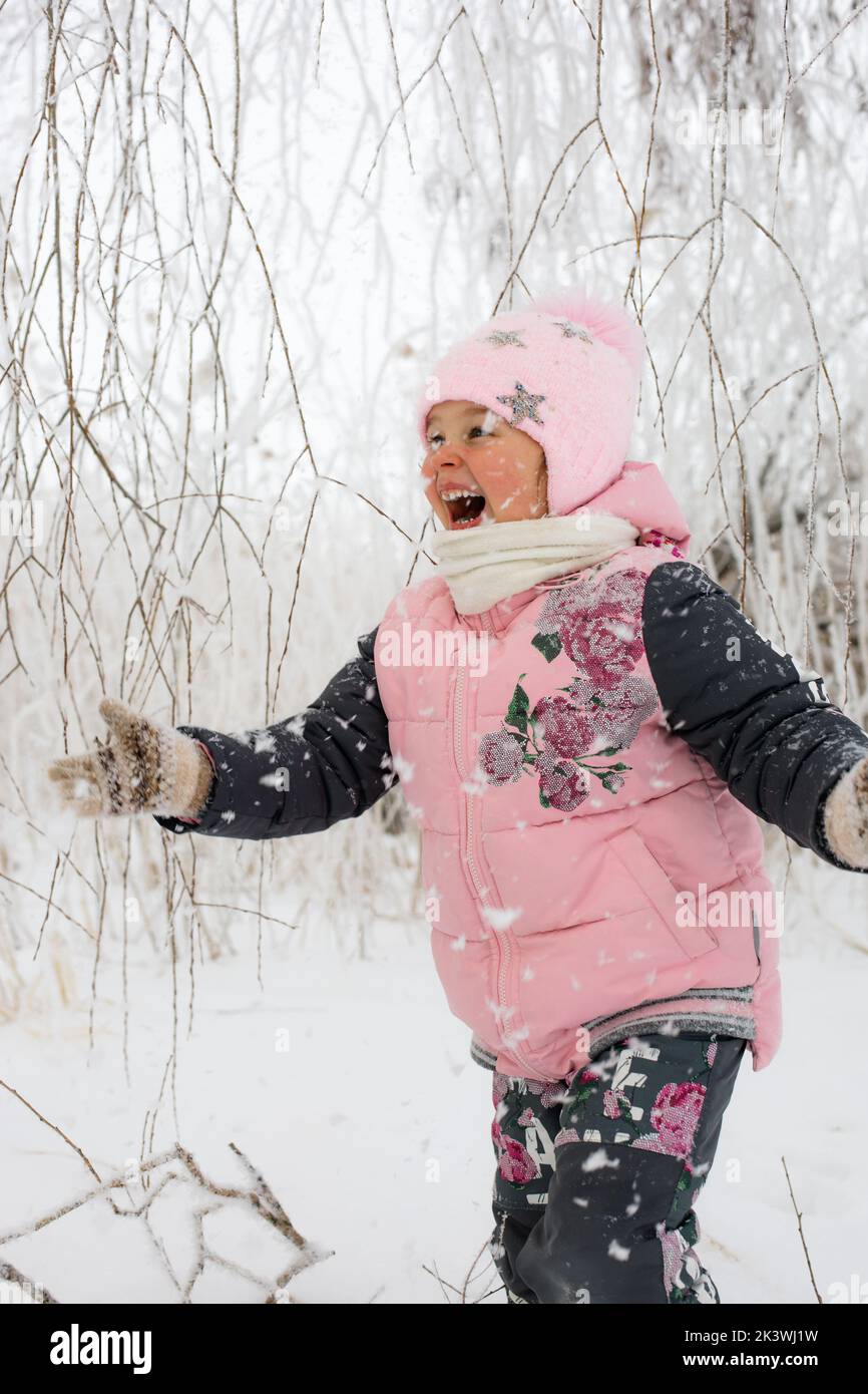 Kleines Mädchen mit ruddigen Wangen und breiten extrem glücklichen Lächeln in warmen Winterkleidung, die auf Schnee mit verschneiten Bäumen im Hintergrund läuft. Winterspaziergang Stockfoto