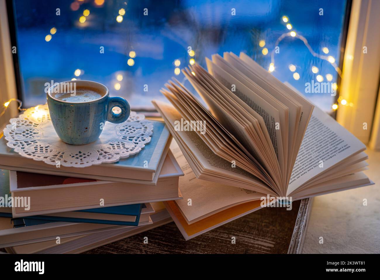 Kaffeetasse, offenes Buch, Lichterketten auf Fensterbank auf blauem unscharfem Hintergrund. Konzept warme und gemütliche Inneneinrichtung, Bücher lesen, Hygge Stockfoto