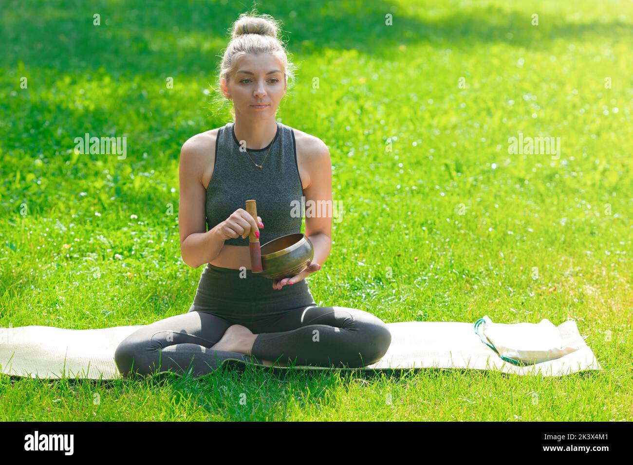 Eine Frau, die im Sommer auf dem grünen Gras im Park sitzt, hält eine Meditationsschale. Stockfoto