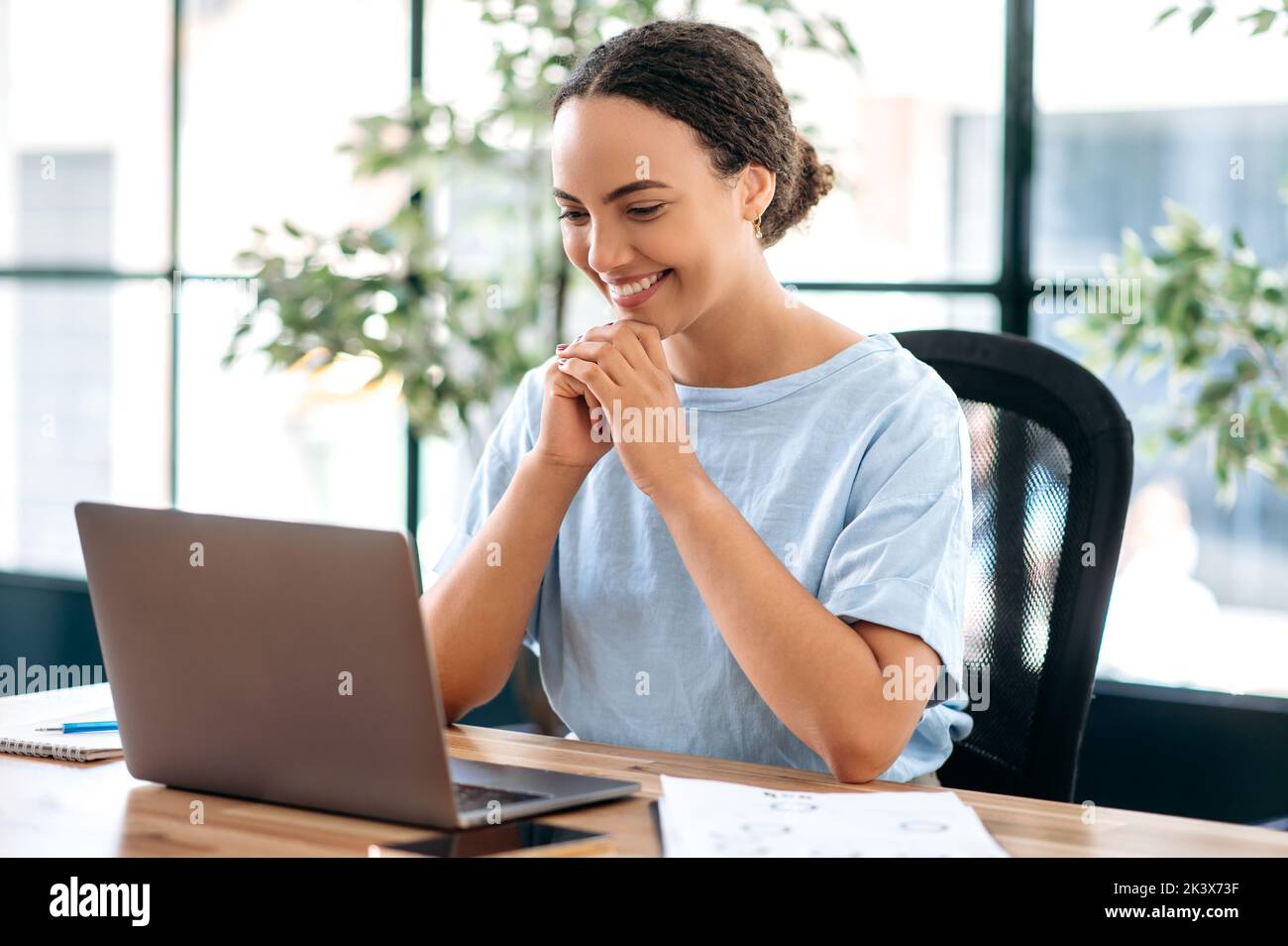 Erfolgreiche selbstbewusste schöne brasilianische oder latino Geschäftsfrau, Büroangestellte, Kreativmanagerin, sitzt am Arbeitsplatz im modernen Büro, schaut auf Laptop, liest gute Nachrichten, lächelt glücklich Stockfoto