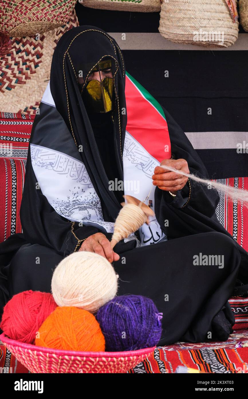 Maskierte arabische Frau aus Abu Dhabi trägt eine Abaya bei einem Folklife Festival, das traditionelle Handwerkskunst demonstriert und Wolle auf eine Spindel setzt. Stockfoto