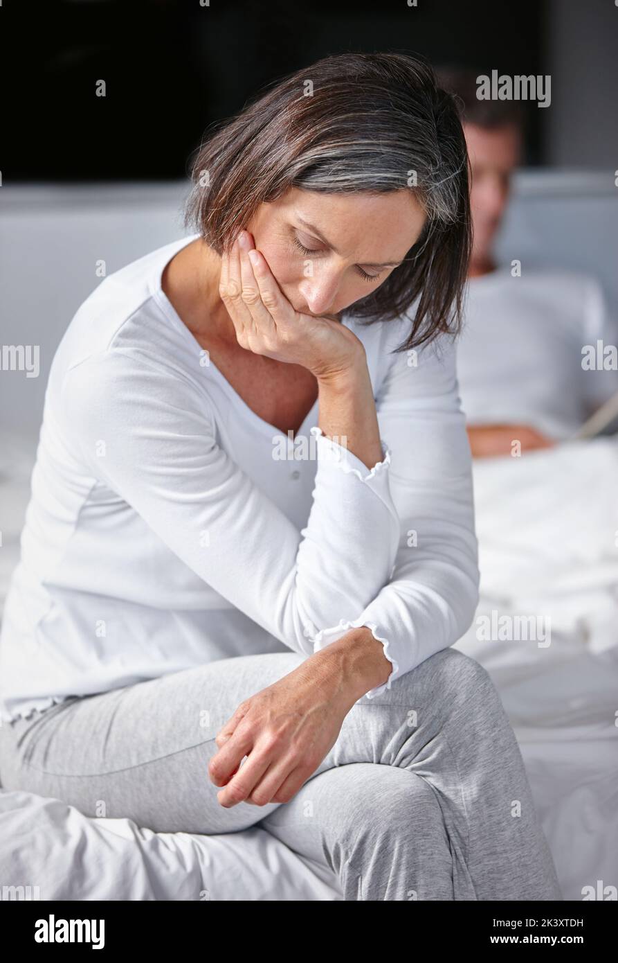 Eine reife Frau, die traurig aussieht, während sie mit ihrem Mann im Hintergrund auf ihrem Bett sitzt. Stockfoto