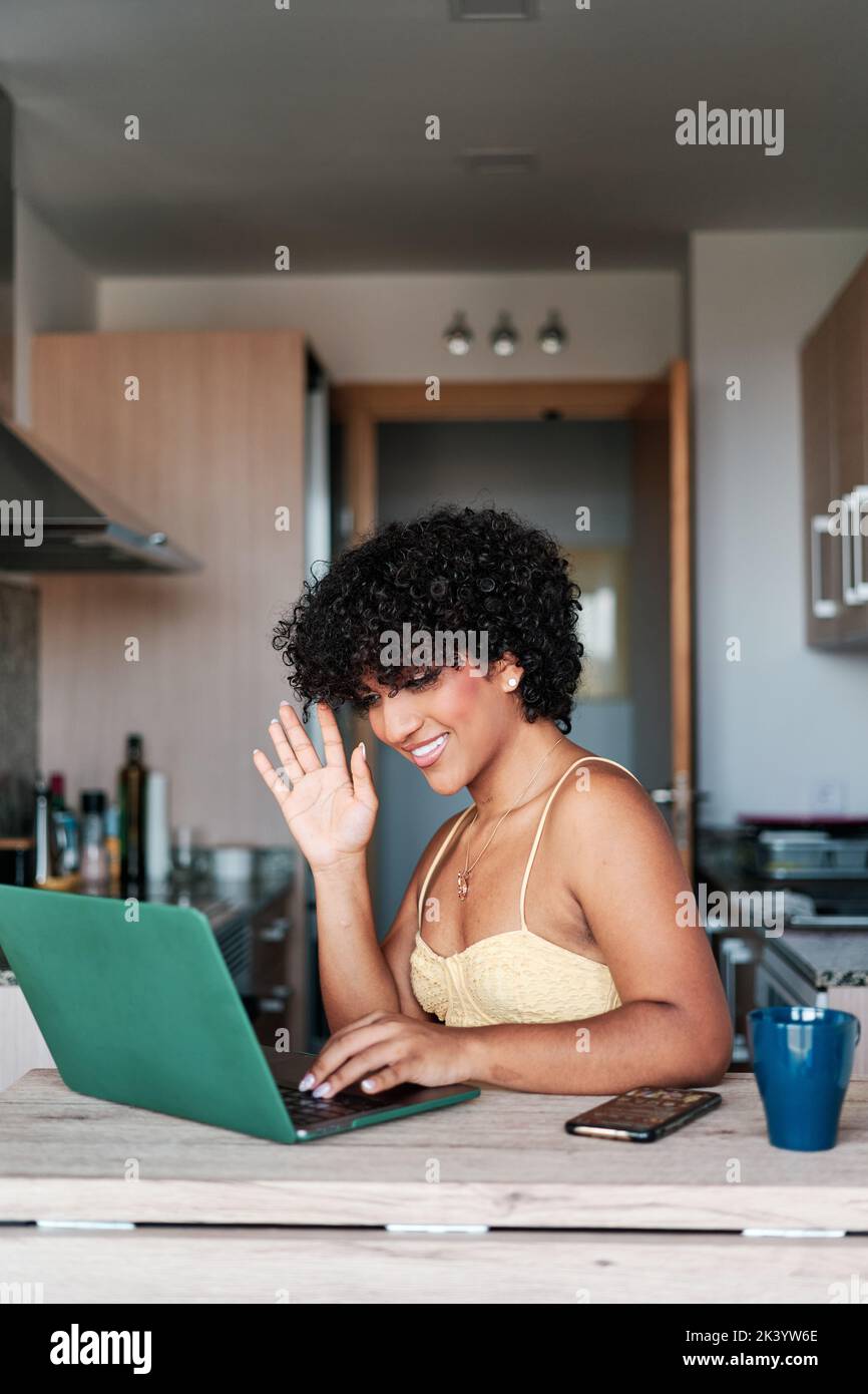 Transgender Frau winkt und lächelt, während sie einen Videoanruf auf einem Laptop in der Küche zu Hause hat. Stockfoto