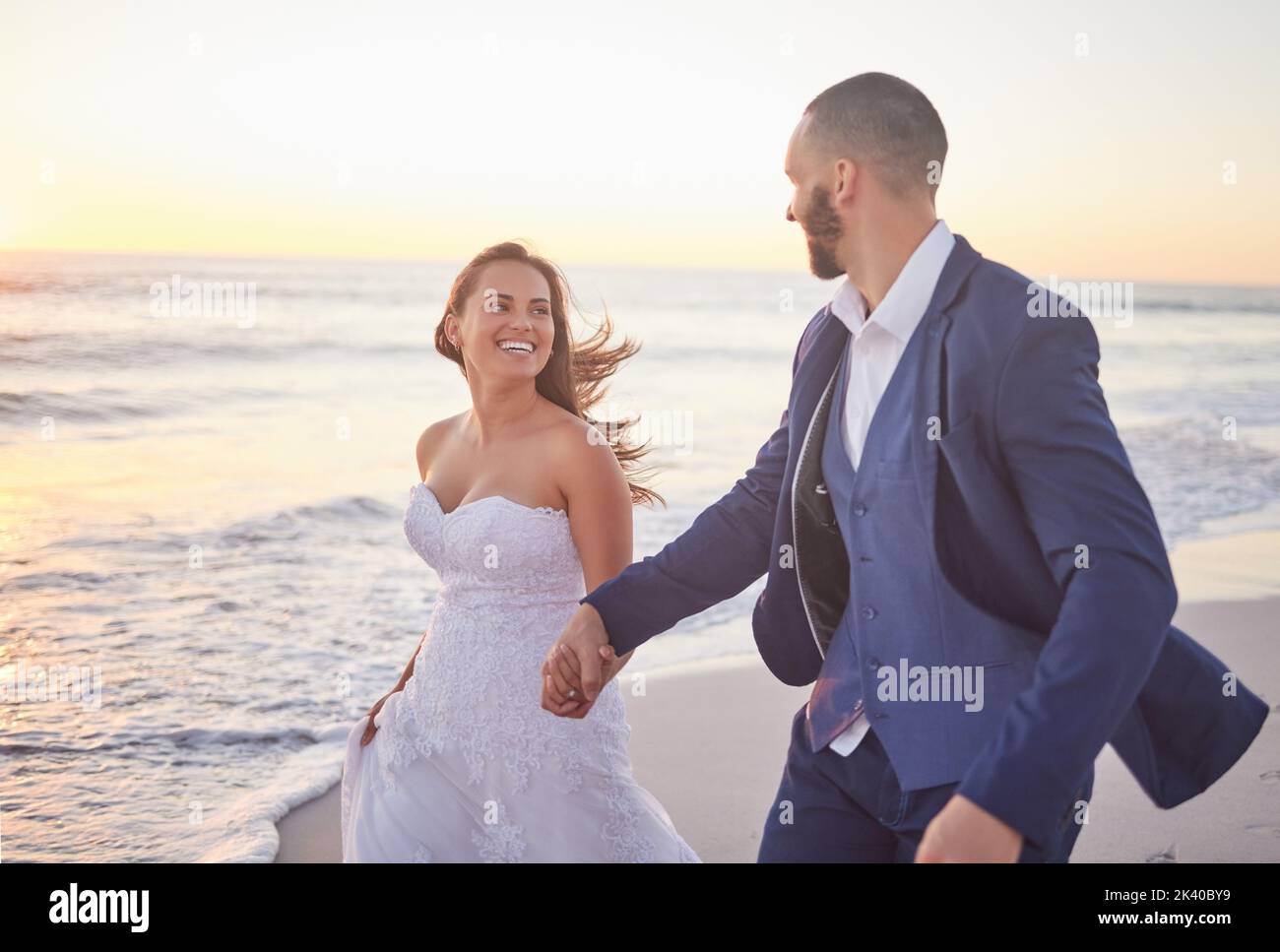 Ehepaar, Hochzeit und Strand lächeln bei Sonnenuntergang auf einem romantischen Spaziergang nach der Hochzeit. Glücklich, Frau und Mann in Anzug zu Fuß am Meer, Romantik und Stockfoto