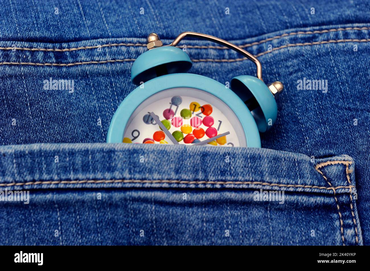 Süßes witziges Young Time Konzept mit einem fröhlichen blauen Wecker mit bunten Bonbons in der Jeanstasche Stockfoto