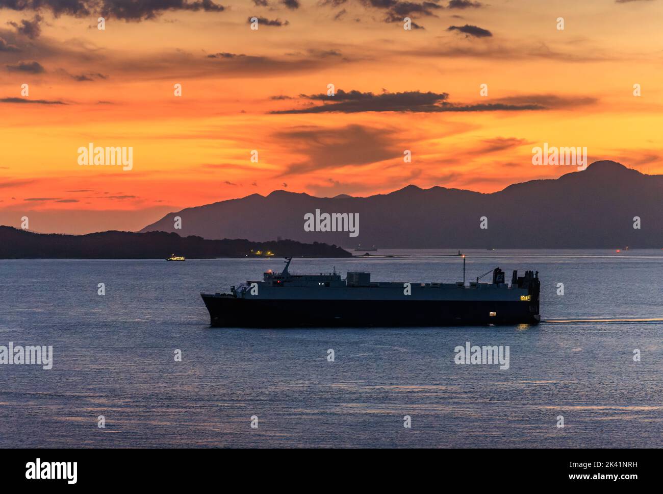 Kleines Containerschiff segelt blaues Meer mit Bergen im Hintergrund bei Sonnenuntergang Stockfoto
