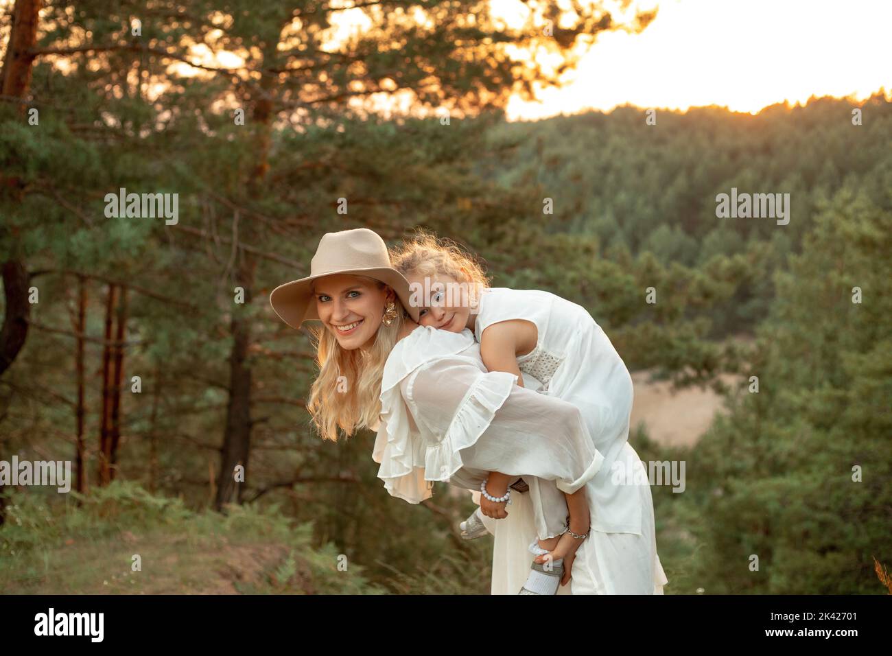 Entzückende, fröhliche, lachende Frau, Mutter im Hut, die ein müdes kleines Mädchen auf dem Rücken trägt, Tochter auf der Wiese im Wald Stockfoto