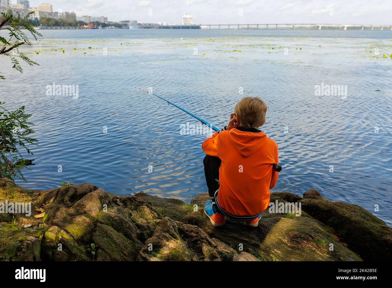 Der Junge sitzt mit dem Rücken und fängt Fische mit einer Angelrute im Fluss Stockfoto