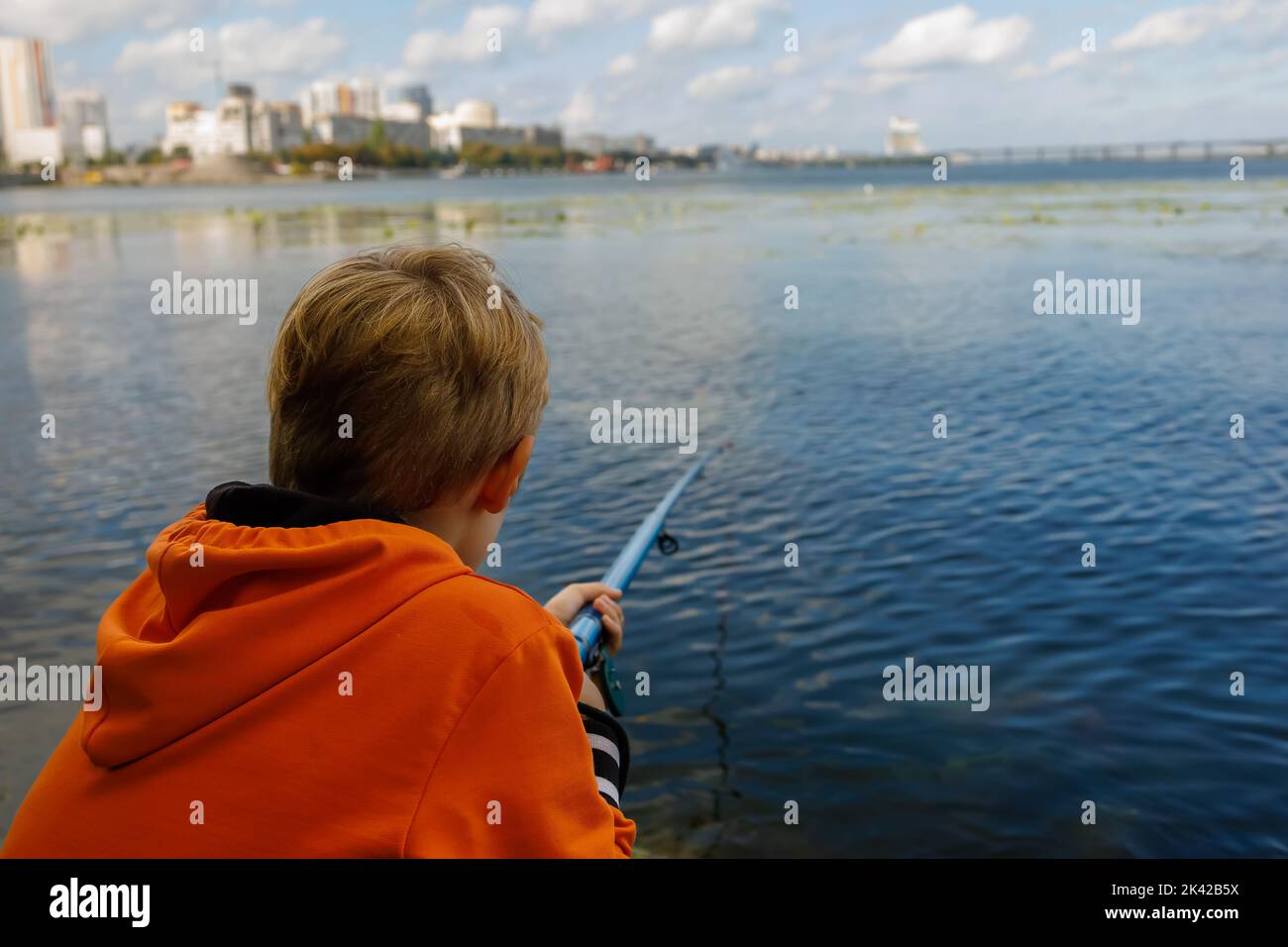 Der Junge fischt mit einer Angelrute am Fluss, hält eine Angelrute in den Händen und sitzt auf den Steinen Stockfoto