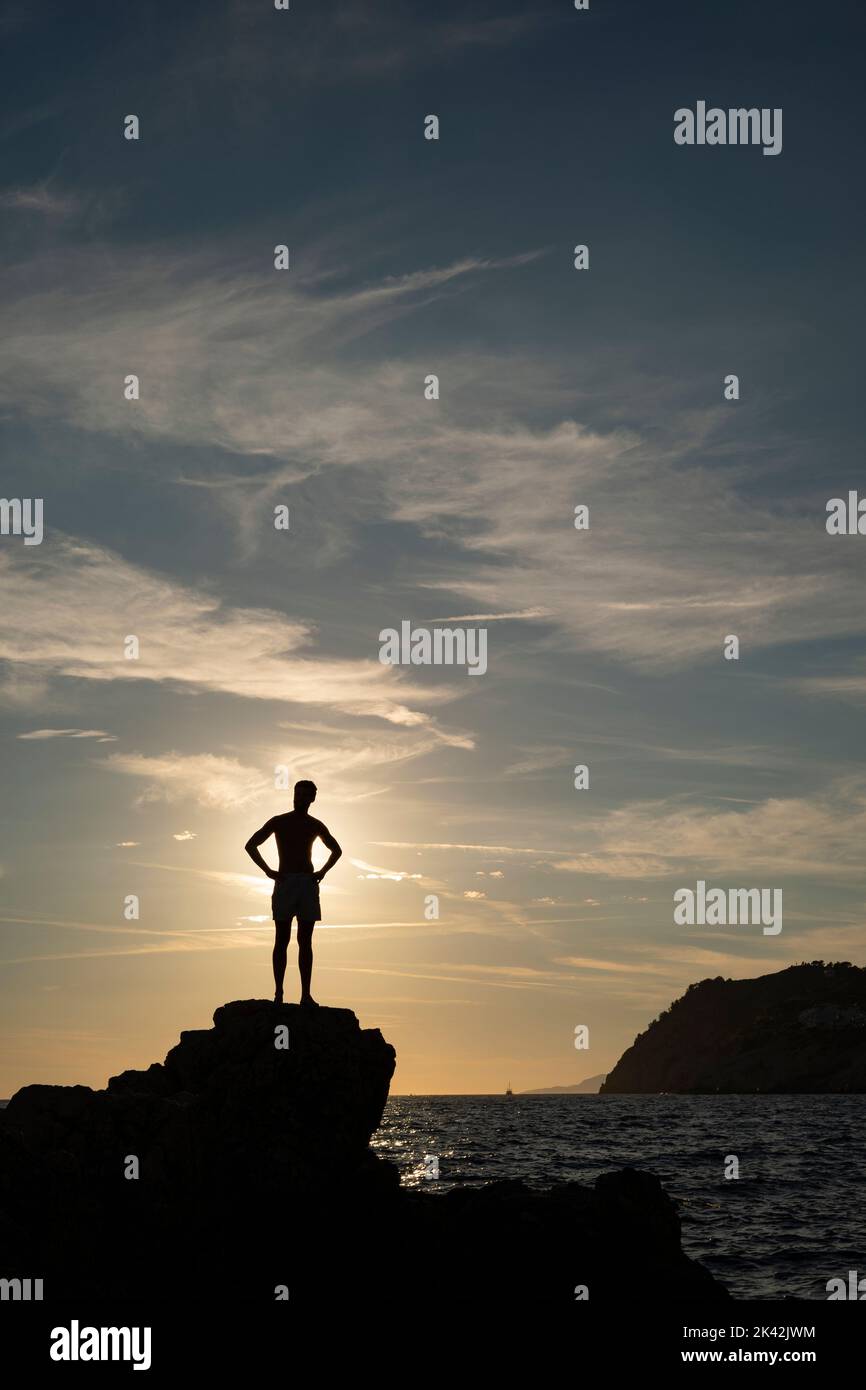 Silhouette eines jungen Mannes, der bei Sonnenuntergang auf einer Klippe am Meer steht Stockfoto