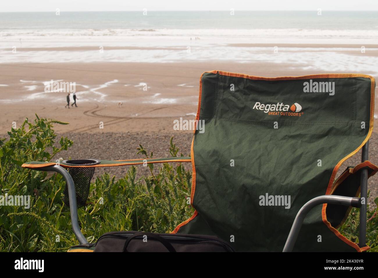 Nahaufnahme eines Campingstuhls mit Blick auf die Widemouth Bay, Cornwall, England, mit der Weite des Sands und zwei Spaziergängern und einem Hund Stockfoto