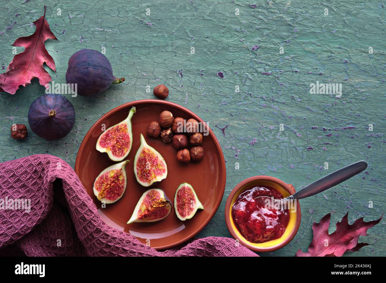 Herbstzeit Hintergrund mit frischen halbierten Feigenfrüchten auf Terrakotta-Keramikplatte. Magentafarbenes Handtuch und rote Herbstblätter auf türkisfarbener, mintgrüner Struktur Stockfoto