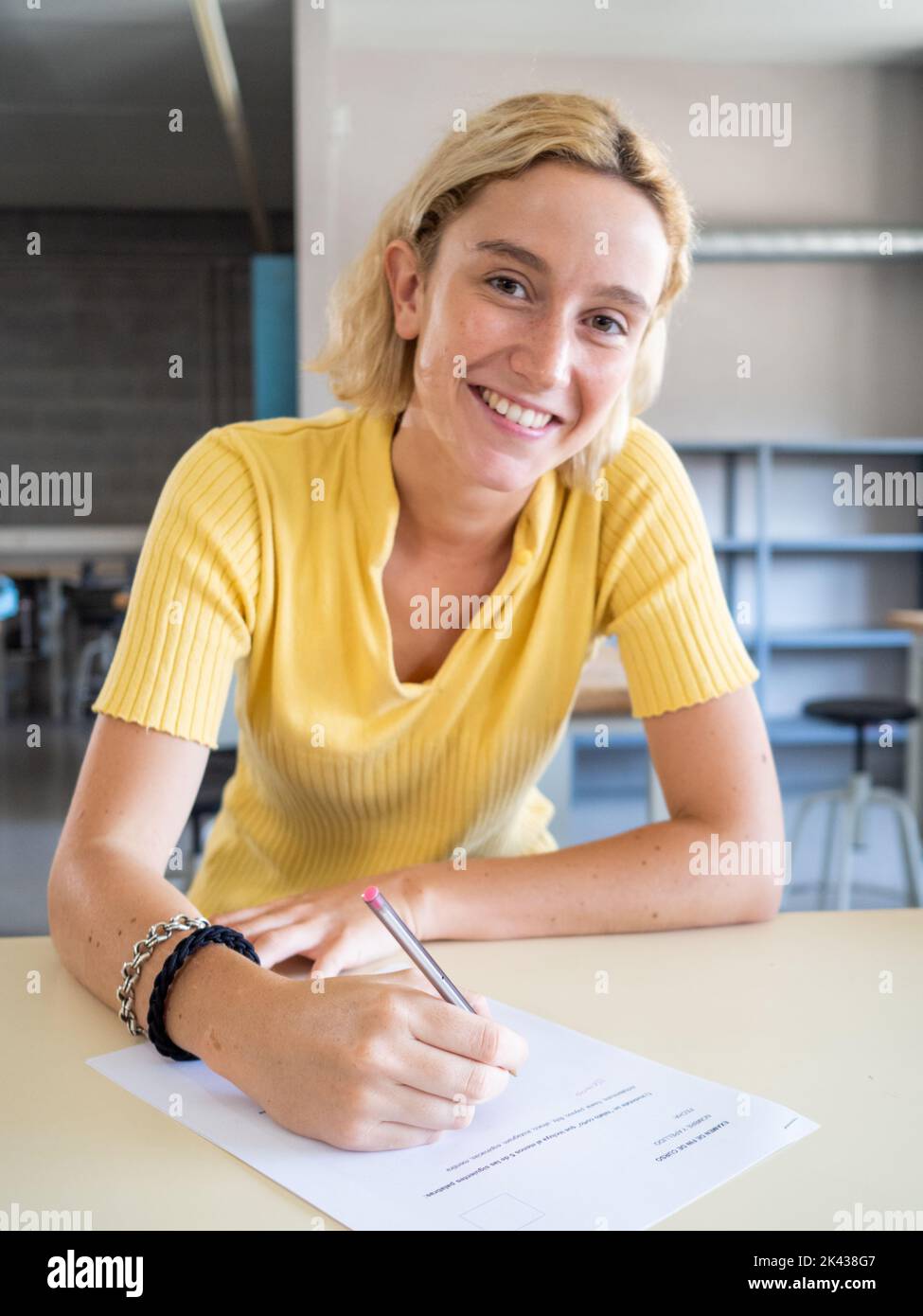Porträt eines lächelnden jungen kaukasischen blonden Schülers, der in einem Klassenzimmer die Kamera anschaut, während er auf Papier schreibt. Bildung, Gymnasium Stockfoto