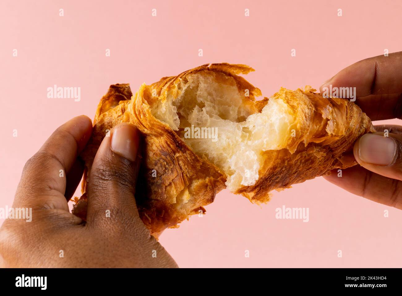 Bild von Händen eines afroamerikanischen Mannes, der Croissants auf rosa Hintergrund zerreißt Stockfoto