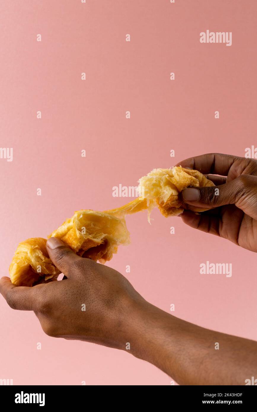 Vertikale Abbildung der Hände eines afroamerikanischen Mannes, der Croissants auf rosa Hintergrund zerreißt Stockfoto
