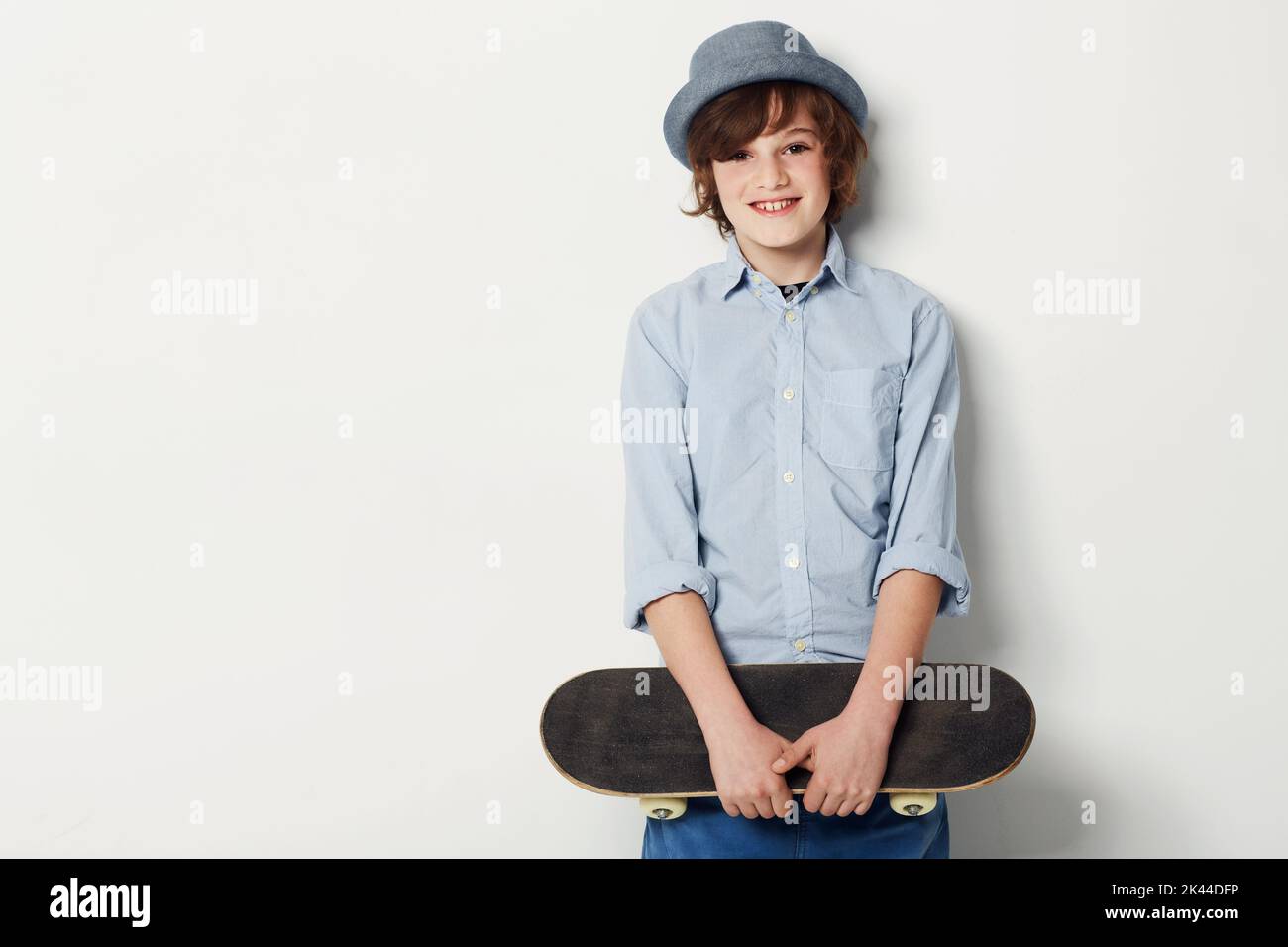 Sein bevorzugtes Transportmittel. Niedlicher Teenager, der trendige Kleidung trägt und ein Skateboard hält, während er auf Weiß isoliert ist. Stockfoto