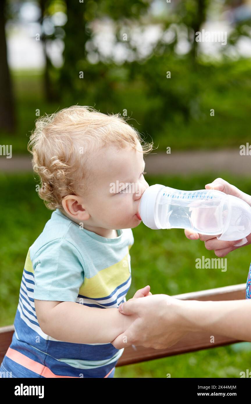 Mutter füttert ihr Baby aus der Flasche mit Wasser im Stadtpark. Die Fürsorge der Mutter für das Kind. Selektiver Fokus, unscharfer Hintergrund Stockfoto