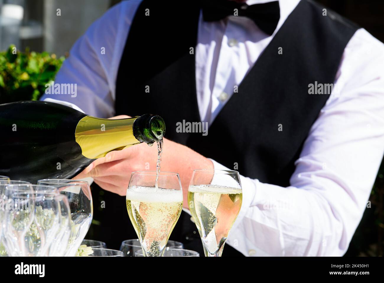 Nahaufnahme einer Champagnerflasche, die Champagner auf einer Sektflöte gegen einen verschwommenen Kellner auf einer Party umschellt Stockfoto