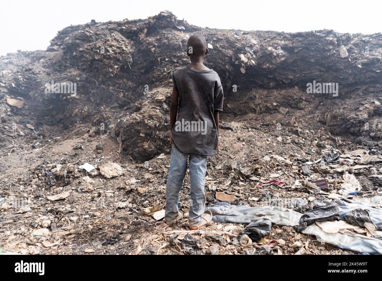 Rückansicht eines jungen afrikanischen Straßenjungen, der in einem städtischen Slum vor einem riesigen Berg rauchenden Mülls steht; Symbol für extreme Armut in den am wenigsten entwickelten Ländern Stockfoto