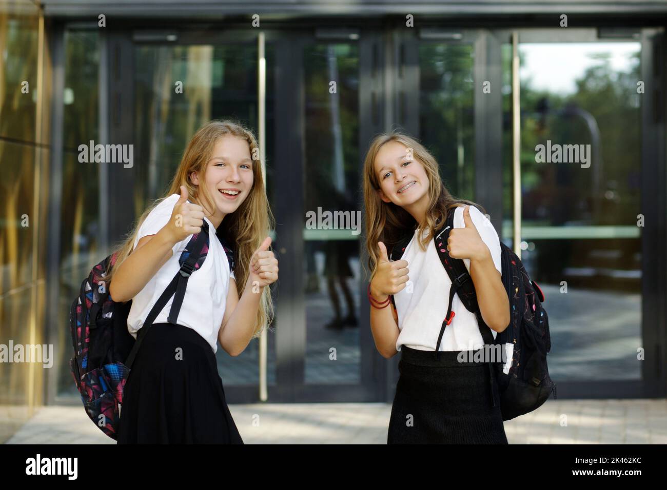 Zurück zur Schule. Erster Schultag. Zwei Mädchen im Teenageralter am Eingang der Schule fühlen sich glücklich und aufgeregt und zeigen die Daumen hoch. Stockfoto