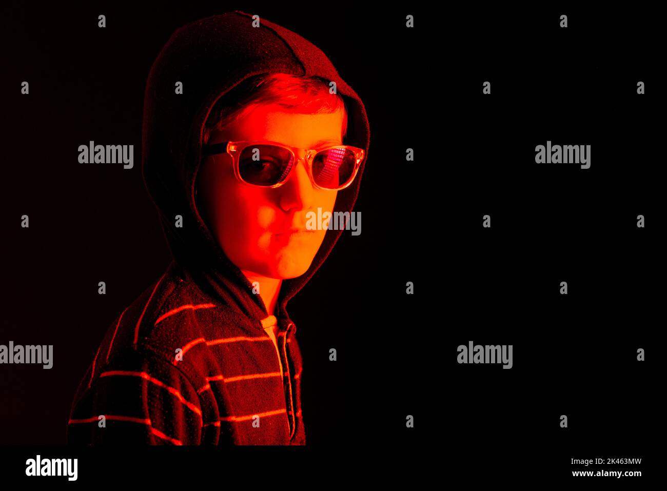 Kaukasisches Kind mit Kapuzen-Sweatshirt und Sonnenbrille, mit ernstem Ausdruck, mit rotem Licht beleuchtet, auf schwarzem Hintergrund und Platz zum Kopieren. Stockfoto
