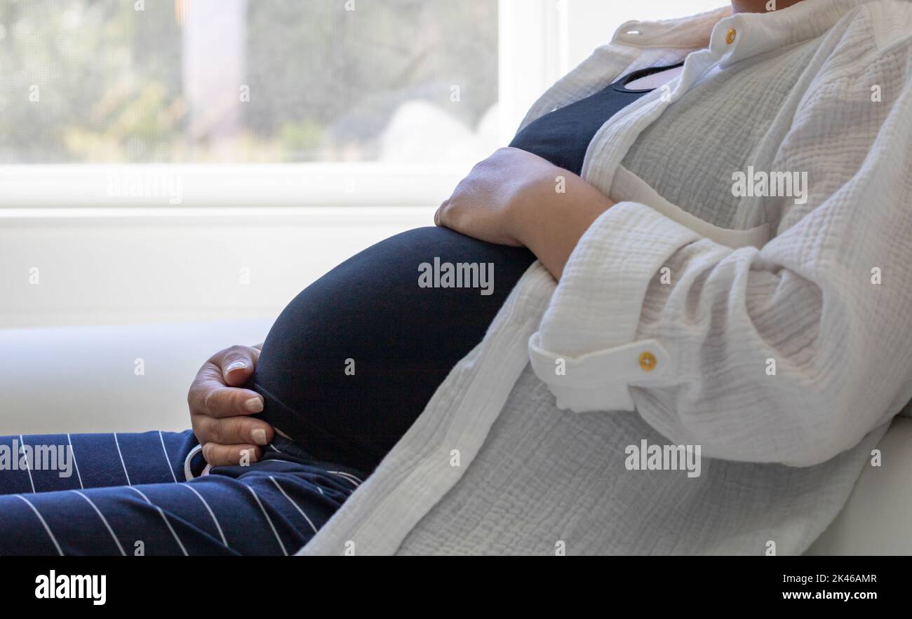Nahaufnahme einer Schwangeren, die auf dem Sofa in der Nähe des Fensters sitzt - Schwangerschaft, Erwartung, Mutterschaftskonzept Stockfoto