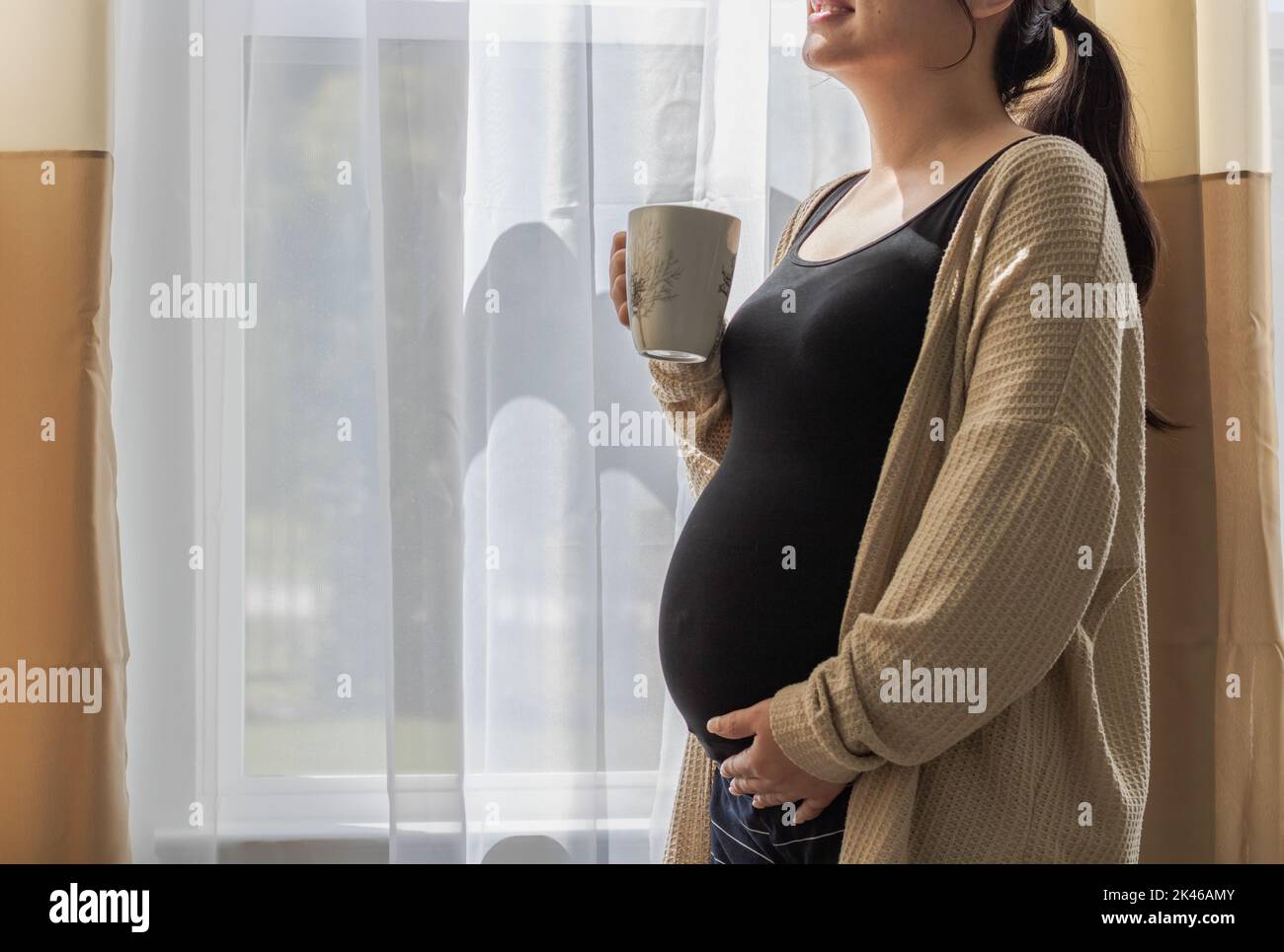 Nahaufnahme einer Schwangeren, die neben dem Fenster mit einem lächelnden Gesicht steht - Schwangerschaft, Erwartung, Mutterschaft Konzept Stockfoto