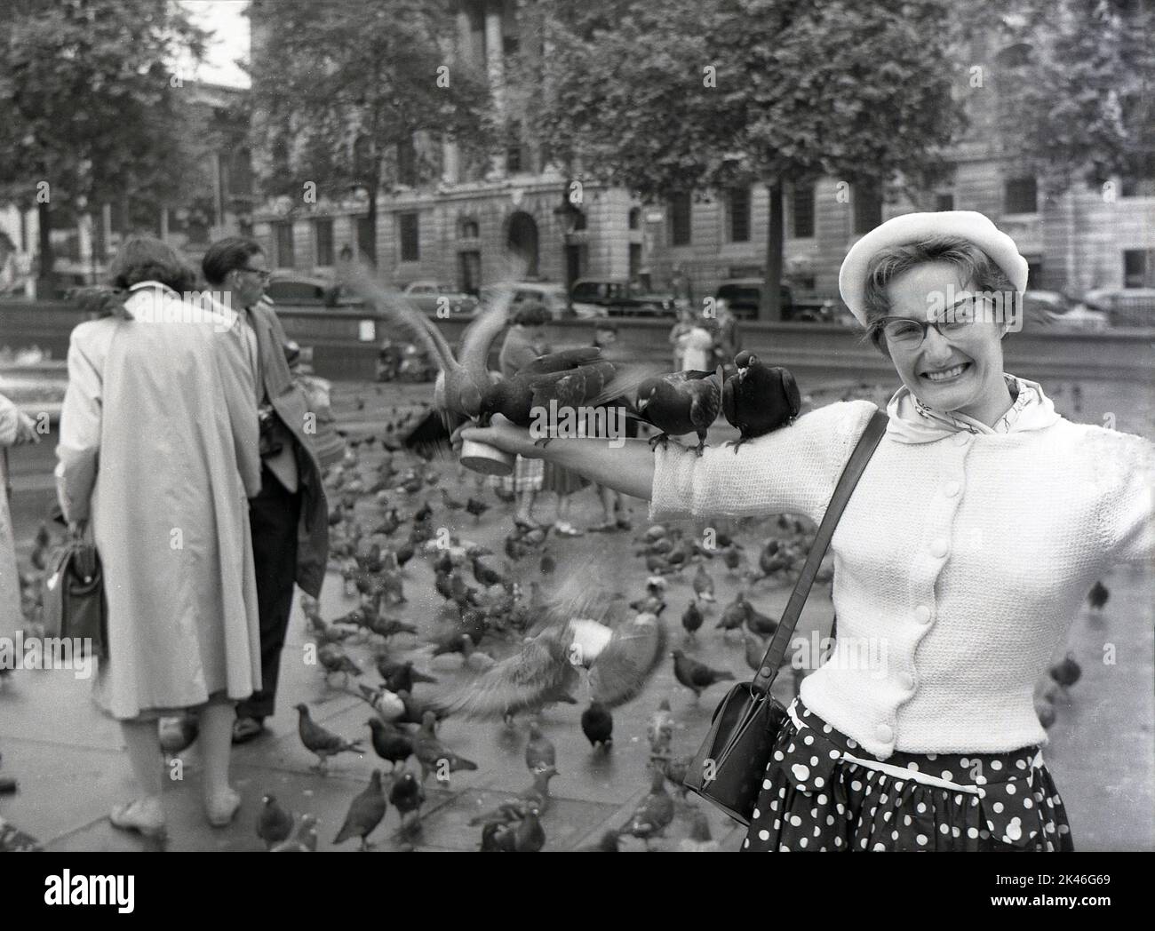 1958, historische Tauben sitzen auf dem ausgestreckten Arm einer aufgeregten Dame am Trafalgar Square, Westminster, London, England, Großbritannien. In ihrer Hand eine kleine Dose Erbsen. Stockfoto