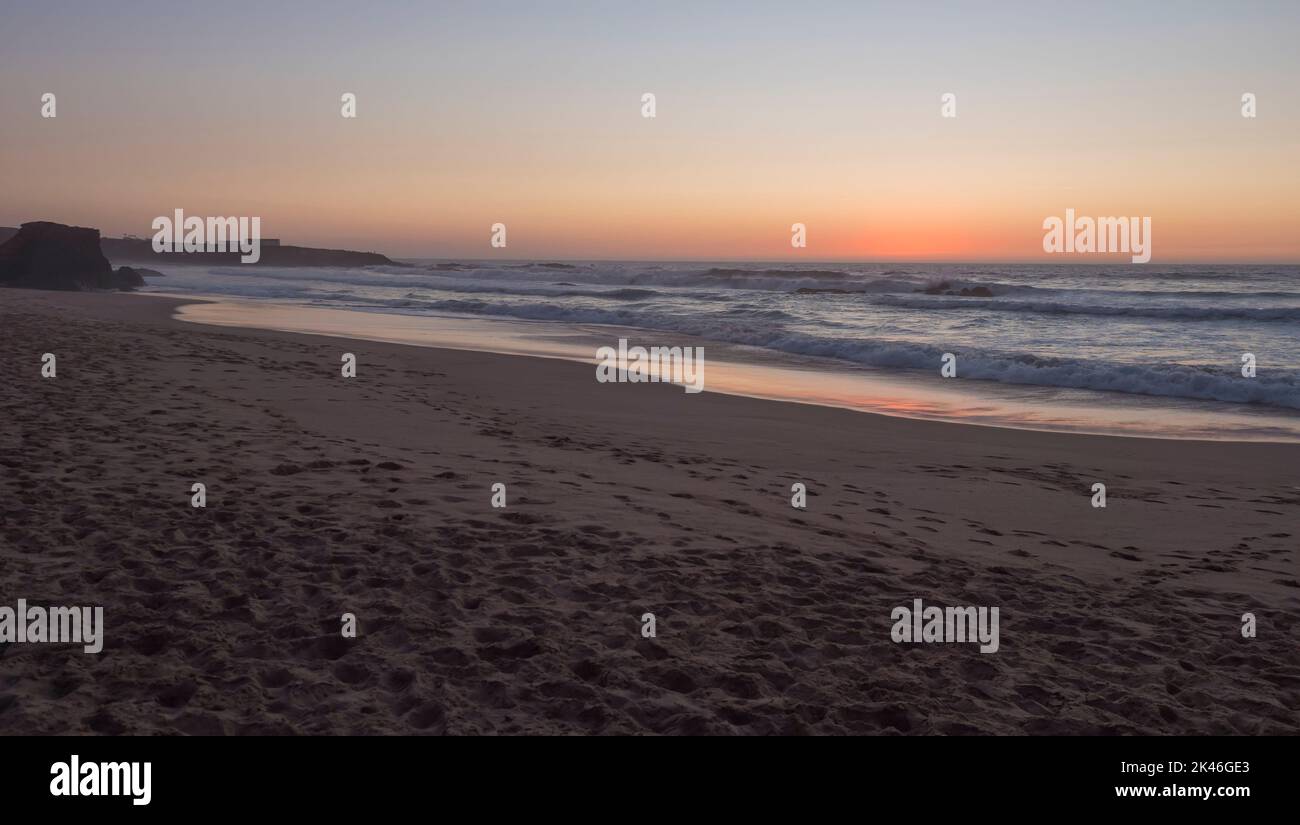 Blick auf den Sandstrand Praia Grande de Almograve bei Sonnenuntergang mit Meereswellen in rosa und rot-blauem Stundenlicht, klarem blauen Himmel. Küste von Rota Vicentina, Almograve Stockfoto