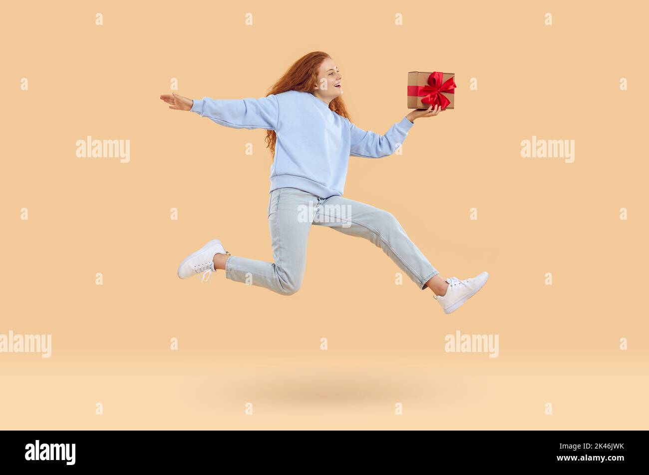 Glückliche junge Frau mit Geschenk-Box springen und fliegen in der Luft auf beigem Hintergrund Stockfoto