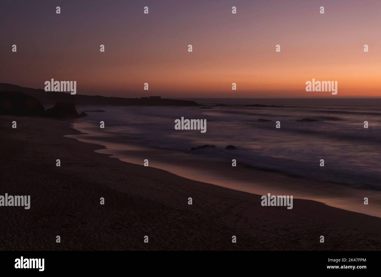 Geheimnisvolle Langzeitansicht des Sandstrandes Praia Grande de Almograve mit verschwommenen Wellen in rosa orange und violettem Licht. Blaue Stunde nach Sonnenuntergang Stockfoto