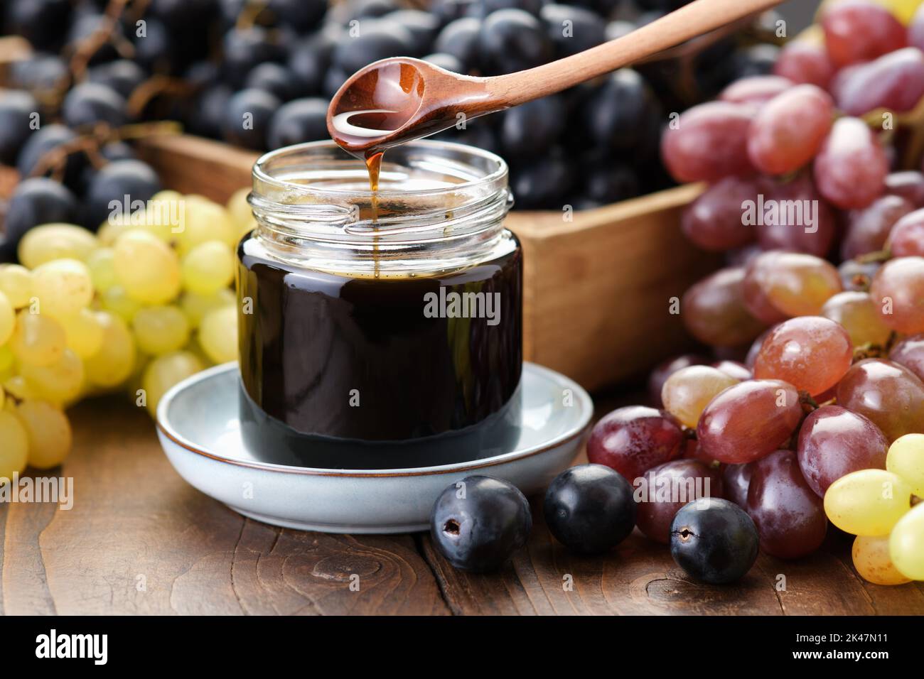Glas aus Traubenmelasse, Traubensirup. Süßstoff, Hustenmedizin. Schwarze, grüne und violette Trauben auf dem Küchentisch. Stockfoto