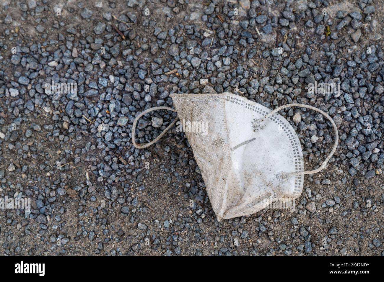 Alte schmutzige weiße Atemschutzmaske, die auf einer Straße aus zerkleinertem Stein liegt. Nahaufnahme von Abfall von einer verlorenen gebrauchten Einweg-Gesichtsmaske, die auf dem Straßenschotter-Gehweg gefallen ist. Stockfoto
