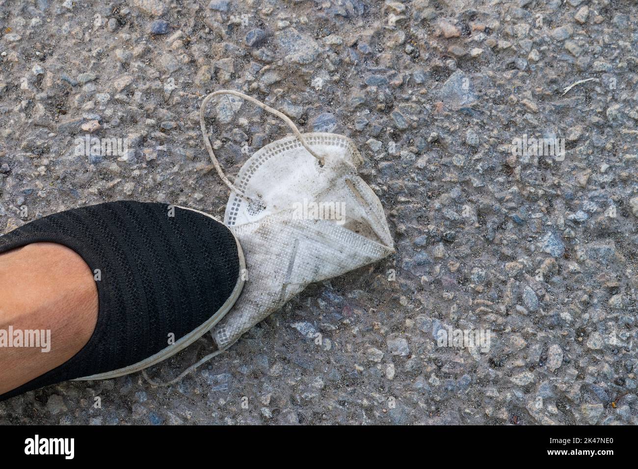 Nahaufnahme eines weißen schmutzigen gebrauchten Atemschutzgeräts mit Fuß in schwarzem Schuh, der auf grauem Bürgersteig steht. Konzept des Endes der Covid-19-Pandemie, Freiheitsbeschränkung. Stockfoto