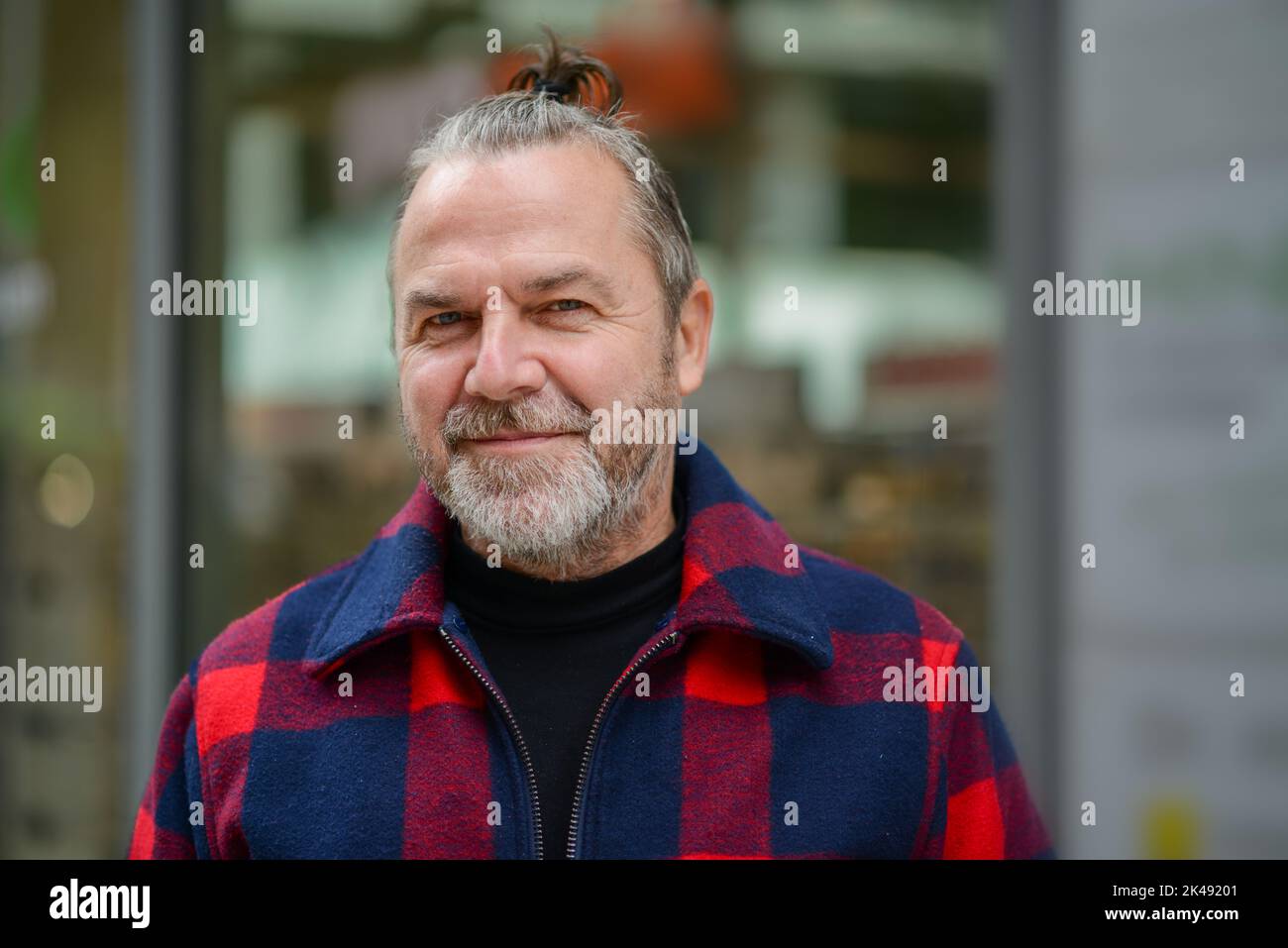 Ein Mann mittleren Alters mit einem chaotischen Brötchen in einer rot-blauen Holzfällerjacke steht mit einem sanften Lächeln auf einer Einkaufsstraße Stockfoto