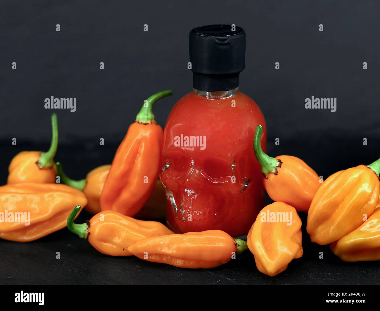 Chilisauce-Flasche in Form eines Schädels zwischen scharfer, orangefarbener Chilischote auf schwarzem Hintergrund Stockfoto