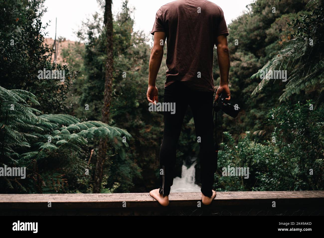 Barfuß kaukasischer Junge in braunem T-Shirt und schwarzer Hose, der auf dem Holzgeländer einer Brücke steht, mit Kamera in der Hand und Blick auf den Waldwasserfall Stockfoto