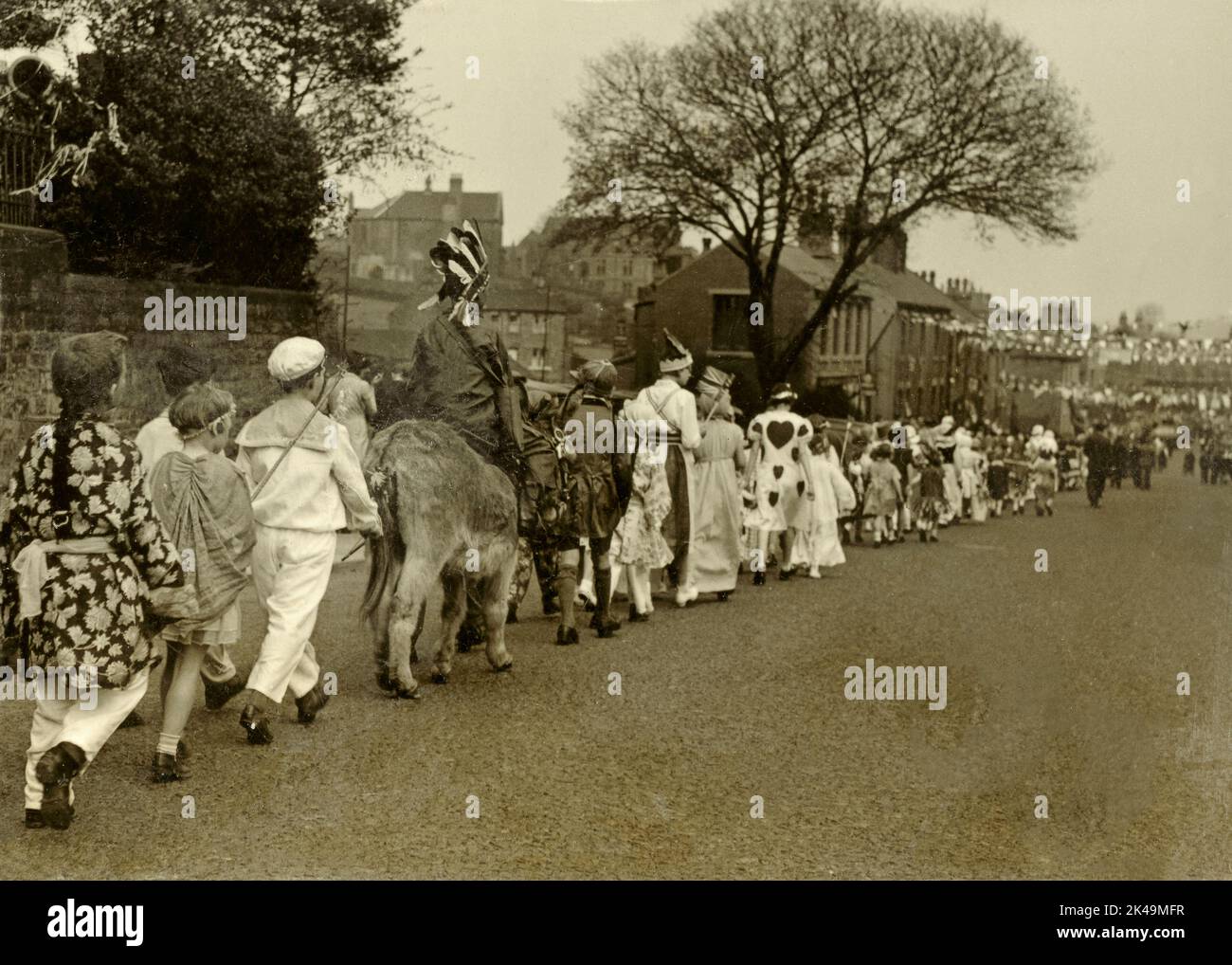 Jugendliche in ausgefallenen Kleidern, darunter ein Junge auf einem Esel, gehen am 6. Mai 1935, dem Silbernen Jubilee-Tag von König George V, in einer langen geraden Straße (wahrscheinlich Wakefield Road) entlang, die mit Fahnen geschmückt ist und sich in Denby Dale in West Yorkshire, England, Großbritannien, befindet. Stockfoto