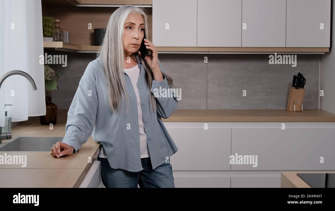Aufgewühlte weiße alte Frau enttäuschte ältere Dame grauhaarige Großmutter Geschäftsfrau steht in der Küche und spricht mit dem Handy unangenehme Anrufe entgegen Stockfoto