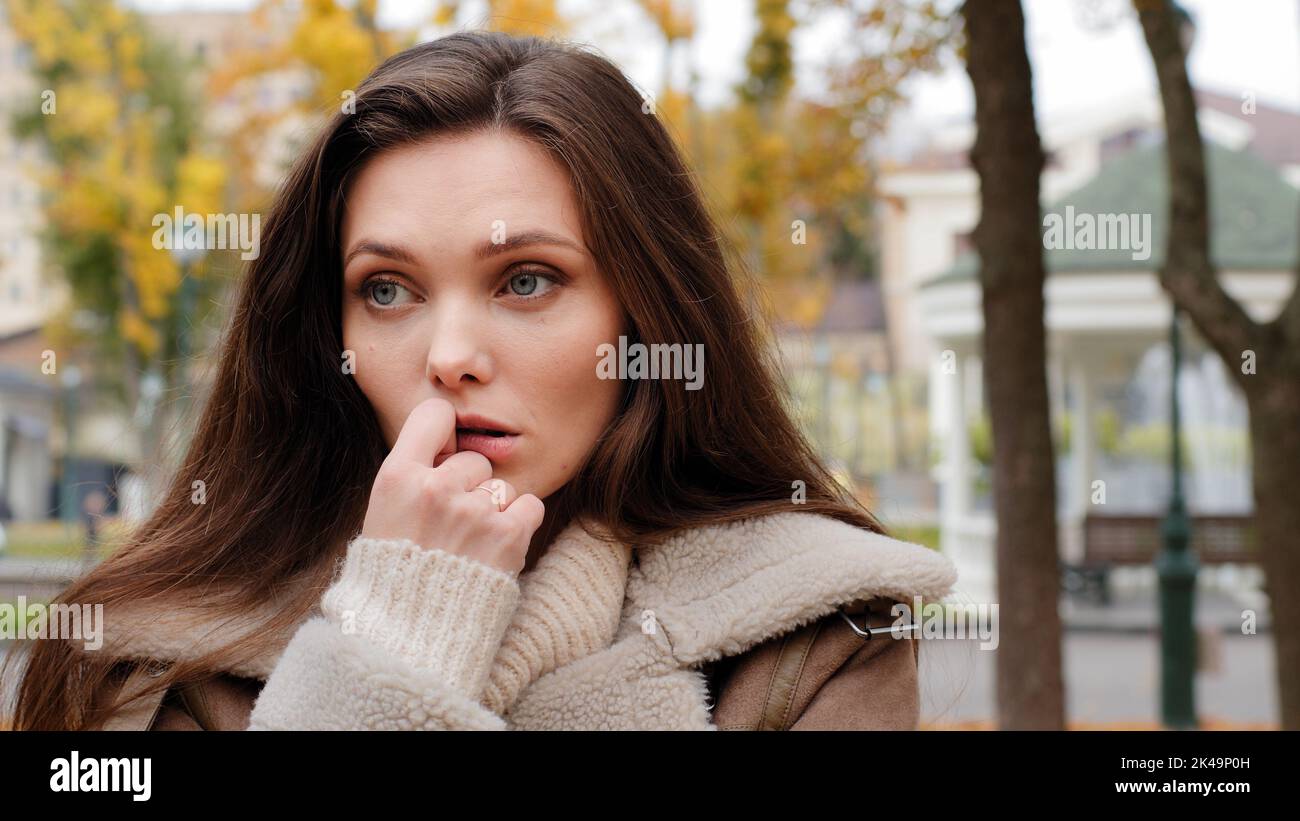 Zweifelhafte junge, brünette, weiße Frau, die allein im Herbst im Stadtpark steht, hält die Hand am Kinn und wartet auf ihren Freund Stockfoto