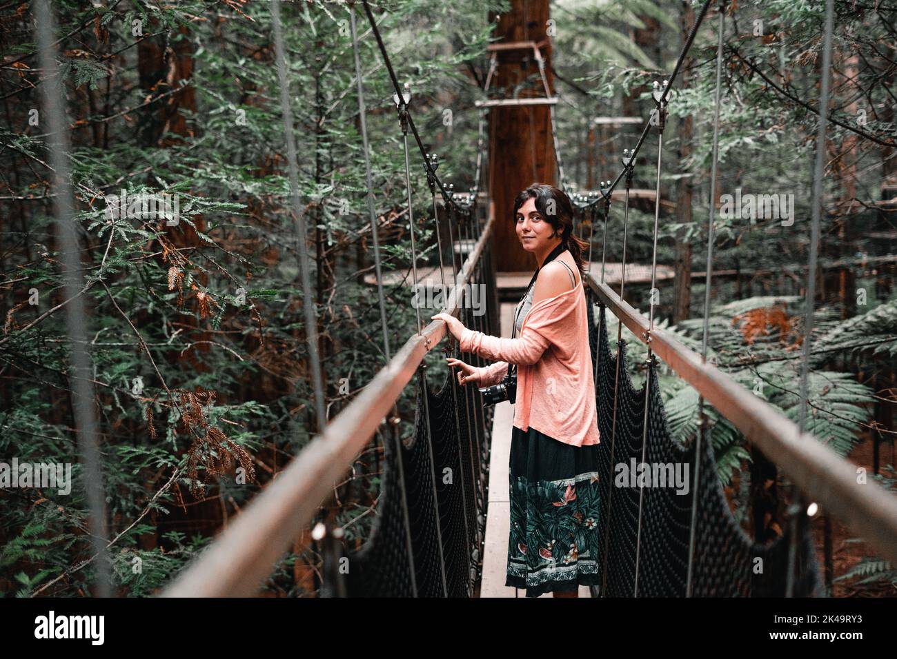 kaukasisches Mädchen mit Kamera um den Hals rosa Bluse und Blumenrock lächeln auf Kamera aus Holzsteg in üppigen Wald, Redwood Baumwalk Stockfoto