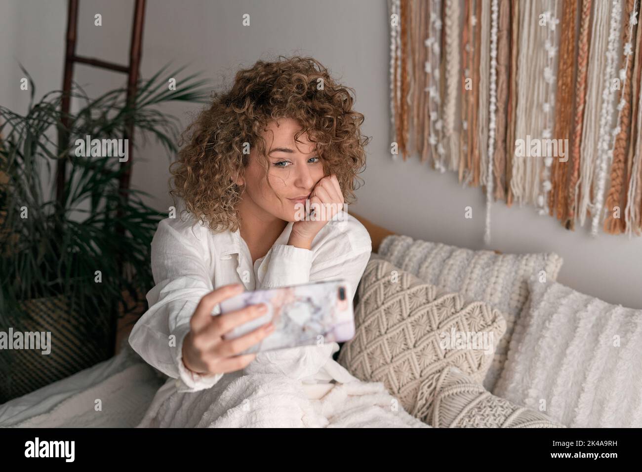 Lächelnde Frau sitzt auf dem Bett und nimmt Selfie auf dem Smartphone Stockfoto