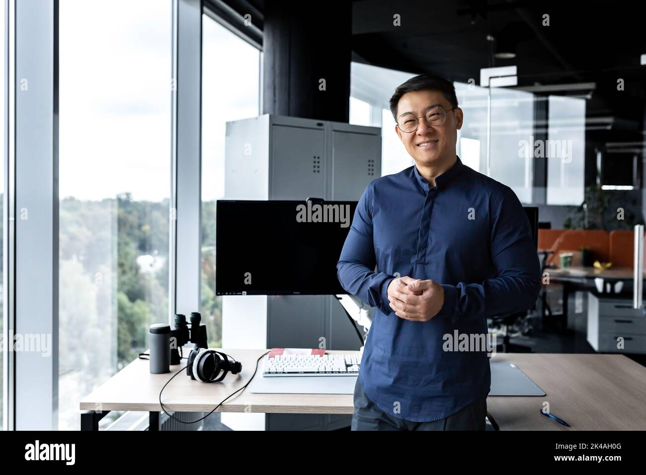 Programmierer in Brille und Hemd lächelnd und schauend auf Kamera, asiatischer Mann, der in einem modernen Computerunternehmen arbeitet, im Büro mit mehreren Computern und Monitoren arbeitet. Stockfoto