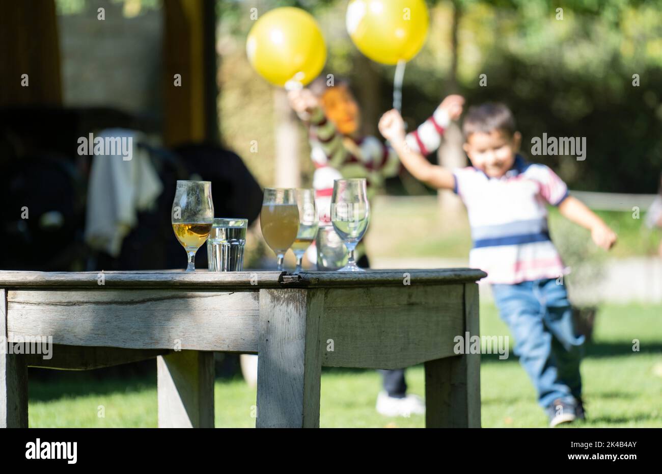 Gläser mit Getränken auf einem Vintage-Tisch während einer Party. Glückliche Kinder laufen mit gelben Ballons auf verschwommenem Hintergrund Stockfoto