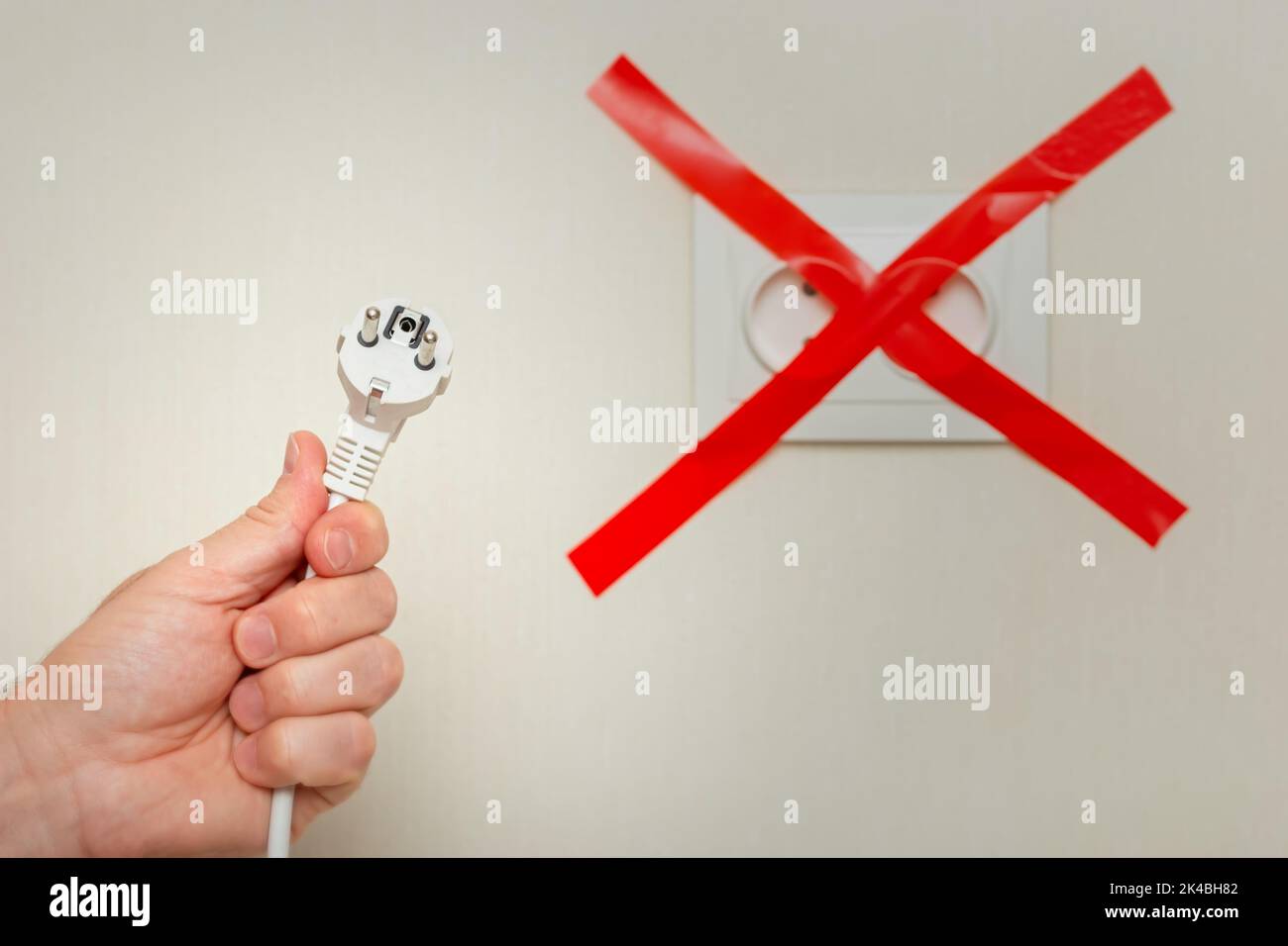 Eine mans Hand hält ein weißes Kabel mit einem Stecker in der Nähe einer Steckdose, das mit einem roten Band quer gebunden ist. Stockfoto