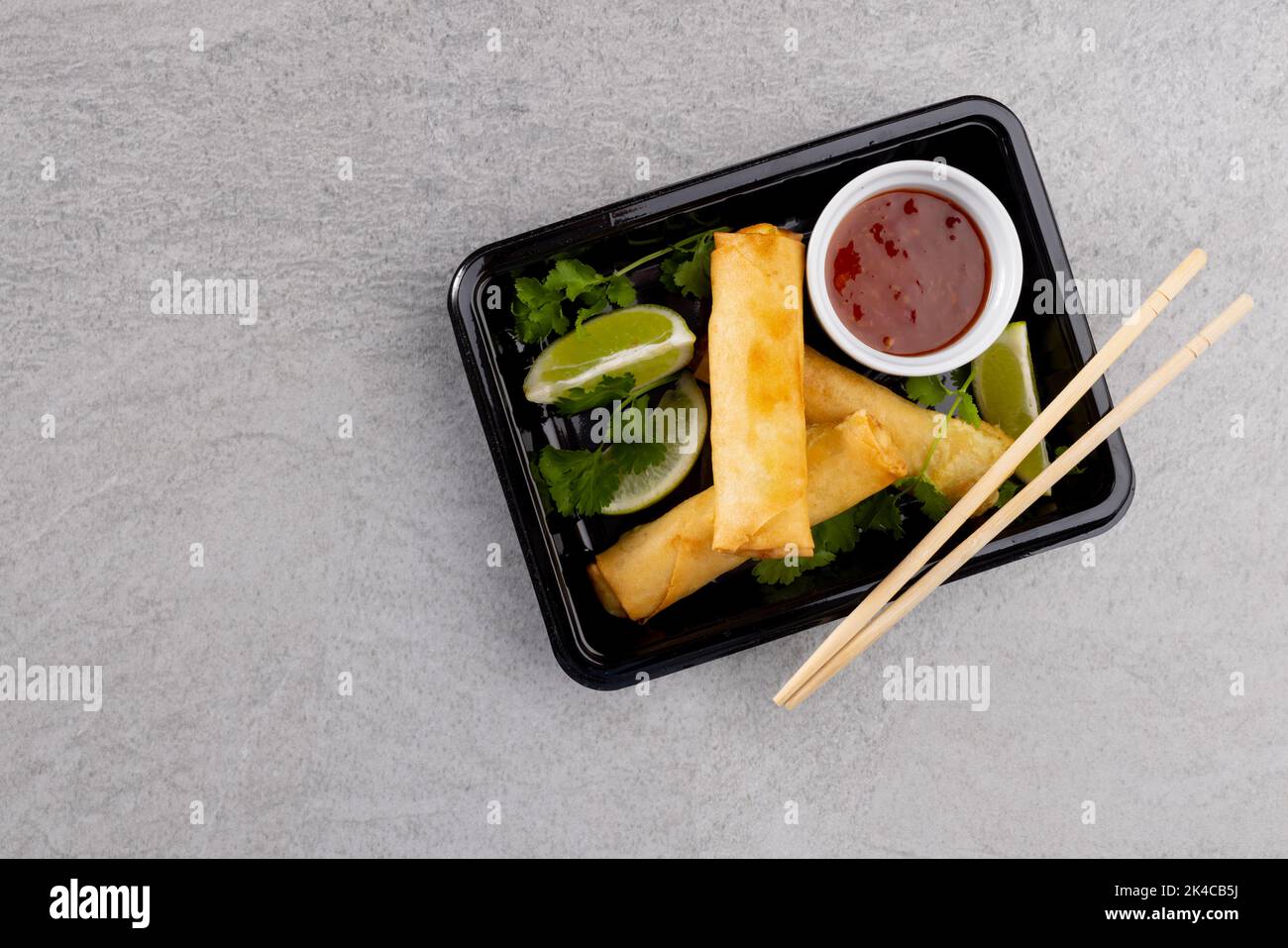 Blick von oben auf asiatische Frühlingsrollen, geschnittene Limette, Chilisauce und Essstäbchen auf grauem Hintergrund Stockfoto