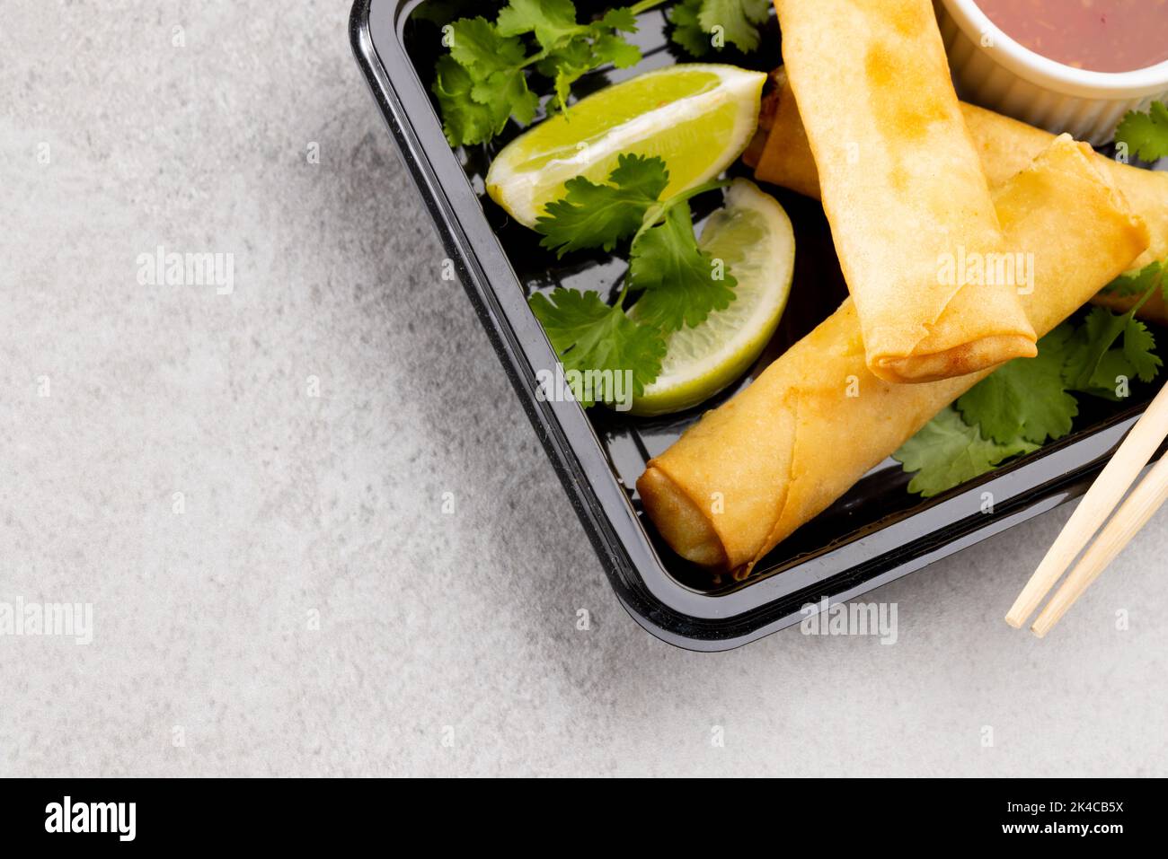 Blick von oben auf asiatische Frühlingsrollen, geschnittene Limette, Chilisauce und Essstäbchen auf grauem Hintergrund Stockfoto